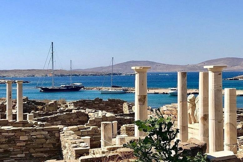 Columns and headless statues on Delos, with sail cruises moored off the site
