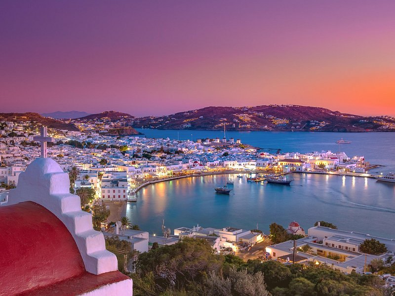 Mykonos Town and the Old Port lit up at dusk, seen over a red-domed chapel